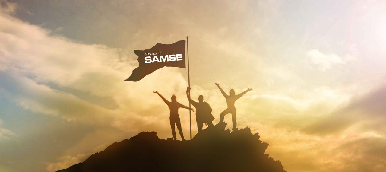 Silhouettes portant un drapeau “Groupe Samse” au sommet d’une montagne au soleil couchant 
