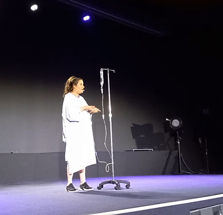 One woman show de l’humoriste Double A. Elle est debout sur scène. Une potence portant une poche pour perfusion est à côté d’elle. 