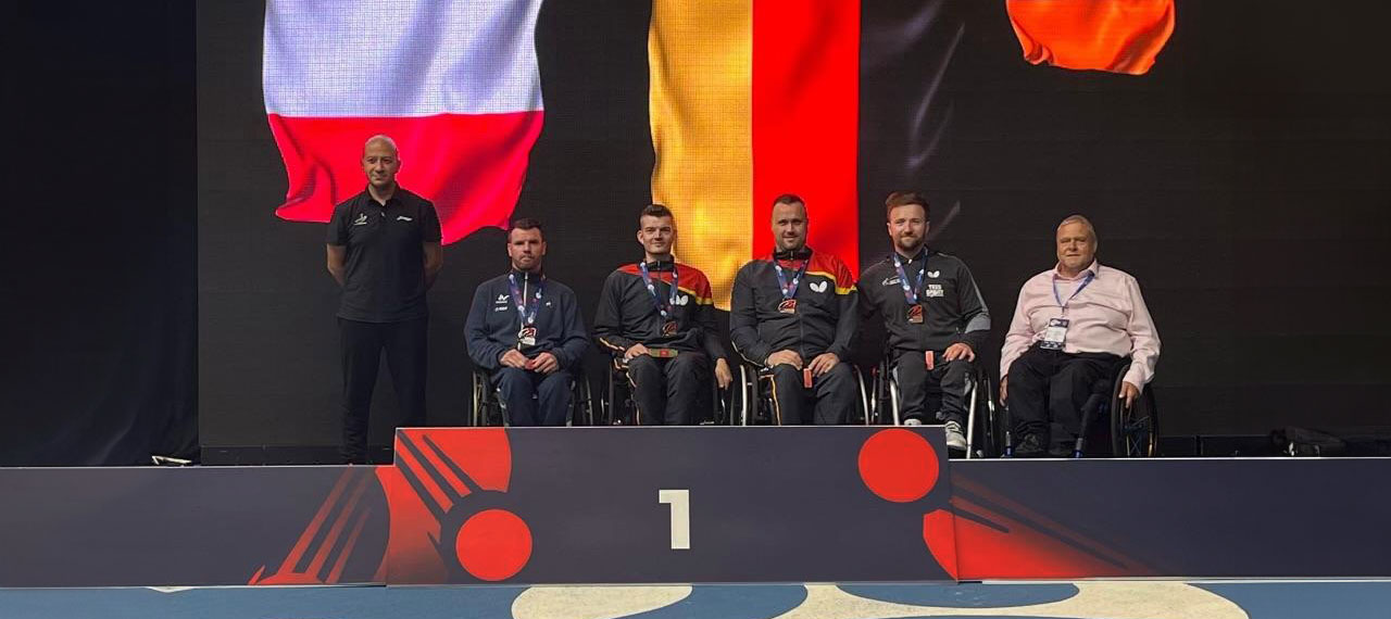 Quatre sportifs en fauteuil roulant sont sur une estrade de remise de prix. Deux entraineurs les accompagnent, l’un d’eux est en fauteuil.