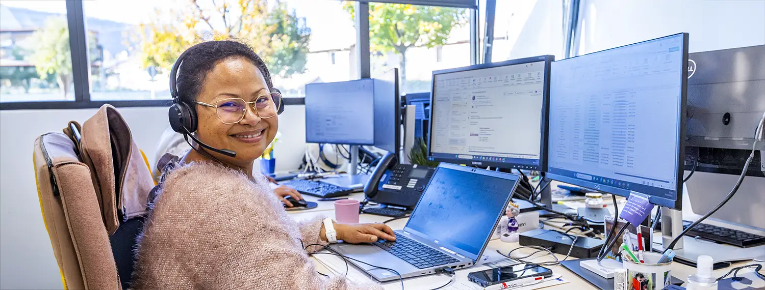 Femme travaillant devant plusieurs écrans, elle est souriante et regarde le spectateur