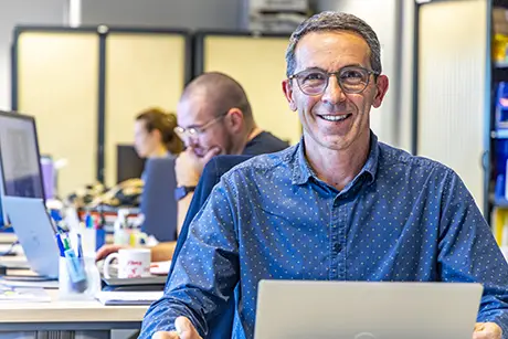 Ambiance de bureau, au premier plan un homme souriant, il regarde le photographe